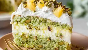 Slice of moist pistachio pineapple cake with whipped topping, crushed pistachios, and pineapple chunks on a plate with a gold fork.