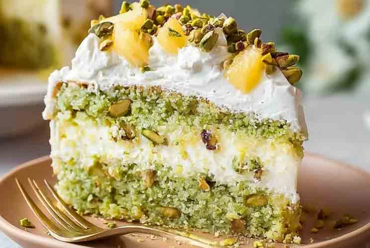 Slice of moist pistachio pineapple cake with whipped topping, crushed pistachios, and pineapple chunks on a plate with a gold fork.