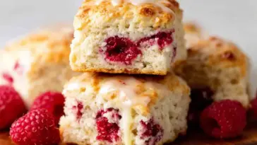 Close-up of fluffy Raspberry Butter Swim Biscuits stacked on a wooden board, filled with baked raspberries and drizzled with vanilla glaze, surrounded by fresh raspberries.