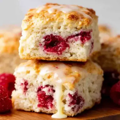 Close-up of fluffy Raspberry Butter Swim Biscuits stacked on a wooden board, filled with baked raspberries and drizzled with vanilla glaze, surrounded by fresh raspberries.