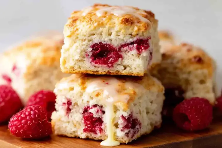 Close-up of fluffy Raspberry Butter Swim Biscuits stacked on a wooden board, filled with baked raspberries and drizzled with vanilla glaze, surrounded by fresh raspberries.