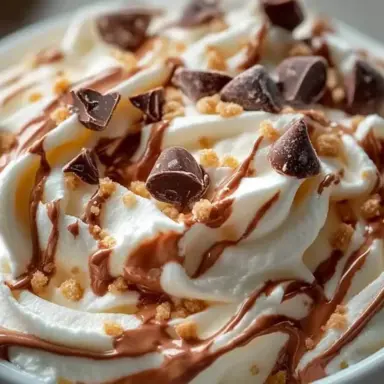Close-up of Reese’s swirl Cool Whip and pudding frosting, topped with chocolate chunks, drizzled peanut butter, and cookie crumbs, showing a creamy, whipped texture.