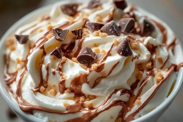 Close-up of Reese’s swirl Cool Whip and pudding frosting, topped with chocolate chunks, drizzled peanut butter, and cookie crumbs, showing a creamy, whipped texture.