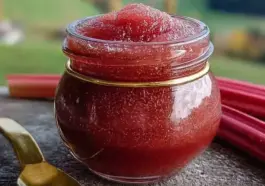 A small round glass jar filled with vibrant red rhubarb butter, topped with a soft swirl of spread, surrounded by fresh rhubarb stalks and a gold spoon, set on a rustic stone surface with a blurred green countryside in the background.