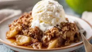 A serving of slow cooker apple crisp topped with a scoop of vanilla ice cream and whipped cream, on a ceramic plate with a fork on the side.