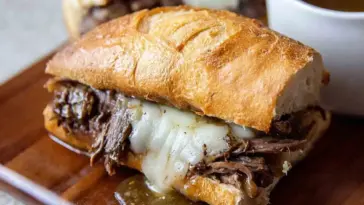 Close-up of a Slow Cooker French Dip sandwich with shredded beef, melted provolone cheese, and golden crusty bread, served with a side of au jus for dipping.