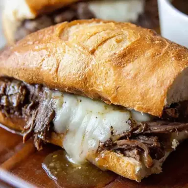 Close-up of a Slow Cooker French Dip sandwich with shredded beef, melted provolone cheese, and golden crusty bread, served with a side of au jus for dipping.