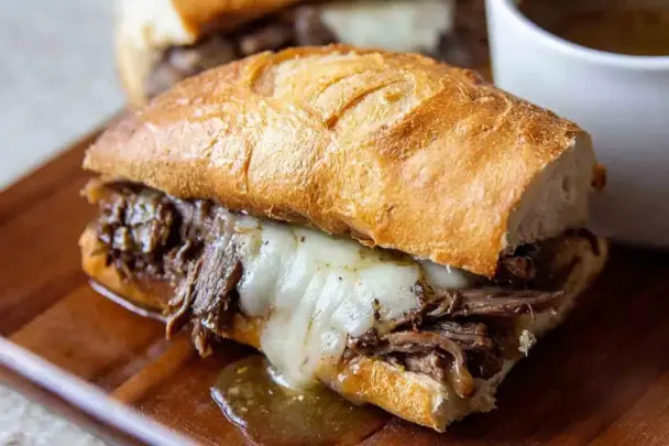 Close-up of a Slow Cooker French Dip sandwich with shredded beef, melted provolone cheese, and golden crusty bread, served with a side of au jus for dipping.