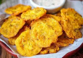 Close-up of crispy Southern Fried Squash slices on a plate lined with parchment paper, with a bowl of dipping sauce in the background.
