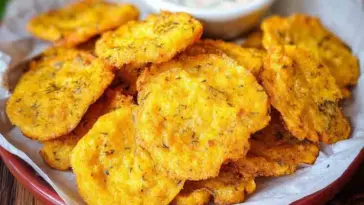 Close-up of crispy Southern Fried Squash slices on a plate lined with parchment paper, with a bowl of dipping sauce in the background.