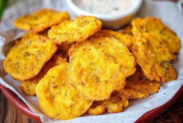 Close-up of crispy Southern Fried Squash slices on a plate lined with parchment paper, with a bowl of dipping sauce in the background.