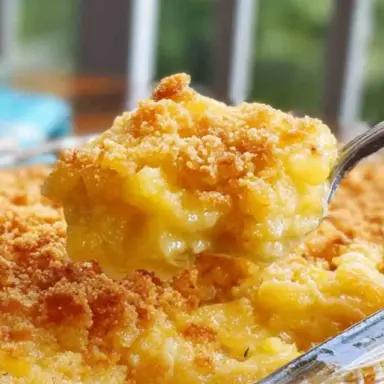 A close-up of a serving spoon lifting a warm, golden-brown Southern Pineapple Casserole from a glass baking dish, showing the creamy pineapple and cheddar filling with a crispy cracker topping.