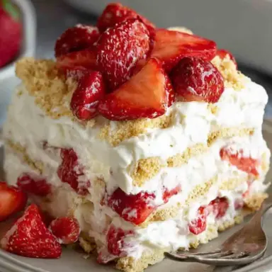 A slice of no-bake strawberry icebox cake with layers of whipped cream, graham crackers, and fresh strawberries, served on a plate with a fork.