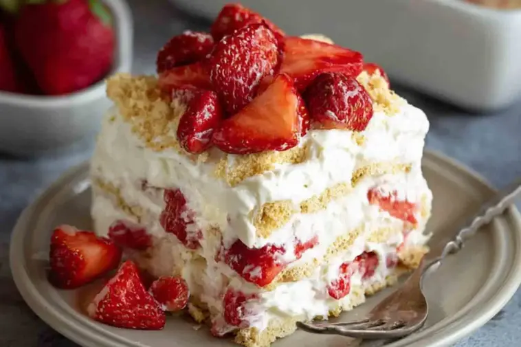 A slice of no-bake strawberry icebox cake with layers of whipped cream, graham crackers, and fresh strawberries, served on a plate with a fork.
