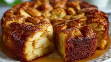 Close-up of a moist Swedish Apple Cake topped with spiced apple chunks, caramel sauce, and powdered sugar, with a slice cut out to reveal its tender crumb.