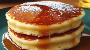 Stack of two fluffy golden pancakes drizzled with maple syrup and dusted with powdered sugar, served on a green plate.