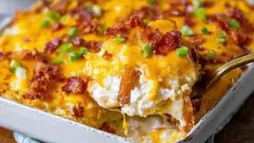 A baking dish filled with cheesy twice baked potato casserole, topped with crispy bacon and green onions, with a serving spoon lifting a hot, gooey portion.