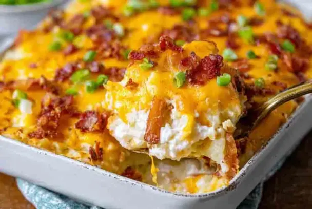 A baking dish filled with cheesy twice baked potato casserole, topped with crispy bacon and green onions, with a serving spoon lifting a hot, gooey portion.