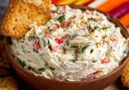 Close-up of a wooden bowl filled with veggie cream cheese topped with chopped red peppers, green herbs, and toasted crackers, with colorful vegetables in the background.