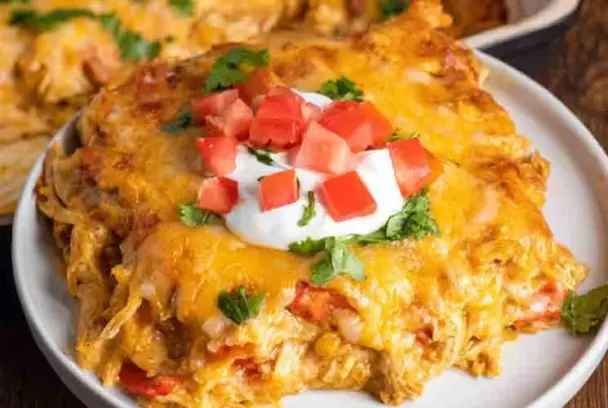 Close-up of a cheesy chicken fajita casserole slice topped with sour cream, diced tomatoes, and chopped cilantro, served on a white plate.