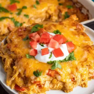 A cheesy chicken fajita casserole slice topped with sour cream, diced tomatoes, and cilantro, served on a white plate with the casserole dish in the background.