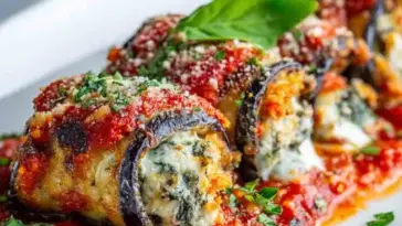 Golden-baked eggplant rollatini filled with spinach and ricotta, topped with marinara sauce, melted cheese, grated parmesan, and garnished with fresh basil on a white plate.