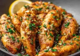 Golden brown garlic butter chicken tenders garnished with chopped parsley, glistening in a skillet with garlic bits and a lemon wedge in the background.