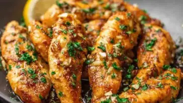 Golden brown garlic butter chicken tenders garnished with chopped parsley, glistening in a skillet with garlic bits and a lemon wedge in the background.