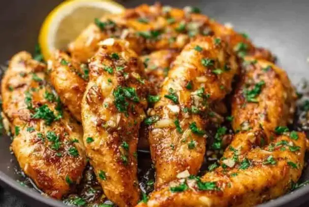 Golden brown garlic butter chicken tenders garnished with chopped parsley, glistening in a skillet with garlic bits and a lemon wedge in the background.