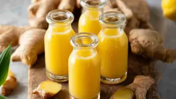 Four glass bottles filled with bright yellow ginger shots placed on a wooden board, surrounded by fresh ginger root and lemon in a cozy kitchen setting.