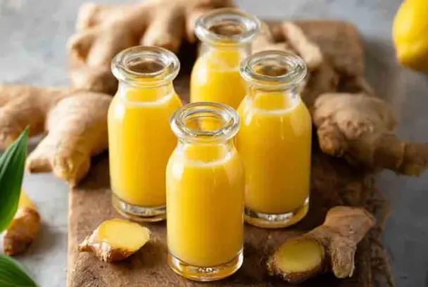 Four glass bottles filled with bright yellow ginger shots placed on a wooden board, surrounded by fresh ginger root and lemon in a cozy kitchen setting.