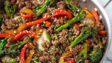 Stir fry made with ground beef, broccoli, red and yellow bell peppers, and sesame seeds in a silver skillet, coated in a savory brown sauce.