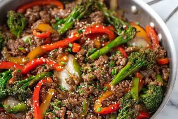 Stir fry made with ground beef, broccoli, red and yellow bell peppers, and sesame seeds in a silver skillet, coated in a savory brown sauce.