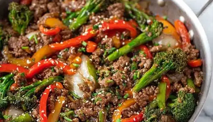 Stir fry made with ground beef, broccoli, red and yellow bell peppers, and sesame seeds in a silver skillet, coated in a savory brown sauce.