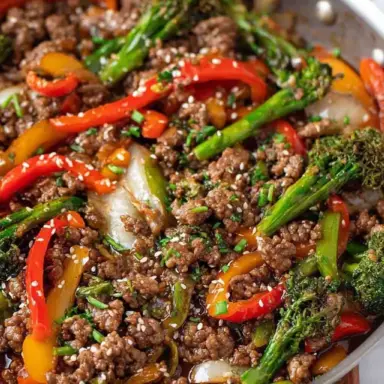 Close-up of a sizzling ground beef stir fry in a skillet with broccoli, red and yellow bell peppers, and sesame seeds, coated in a glossy stir fry sauce.