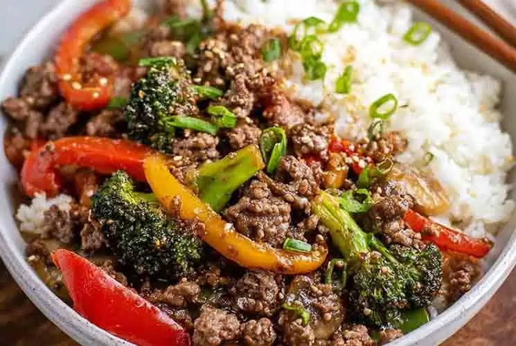 Bowl of ground beef stir fry served over steamed white rice, featuring red and yellow bell peppers, broccoli, sesame seeds, and chopped green onions.