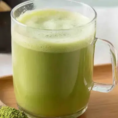 A clear glass mug filled with a vibrant green Japanese Mounjaro weight loss drink made with matcha, apple cider vinegar, and yuzu, sitting on a wooden tray beside a spoonful of green matcha powder.
