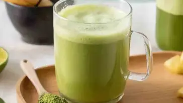 A glass mug of Japanese Mounjaro weight loss drink made with matcha and citrus, resting on a wooden tray beside a spoonful of matcha powder, fresh lime, and ginger in the background.