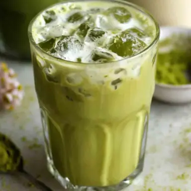 A glass of low calorie iced matcha latte filled with ice cubes and creamy green tea, surrounded by matcha powder and a spoon on a white surface.