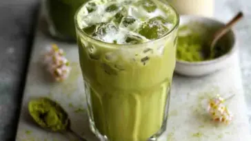 Glass of iced matcha latte made with almond milk and matcha powder, served on a white board with matcha spoon and spring flowers.