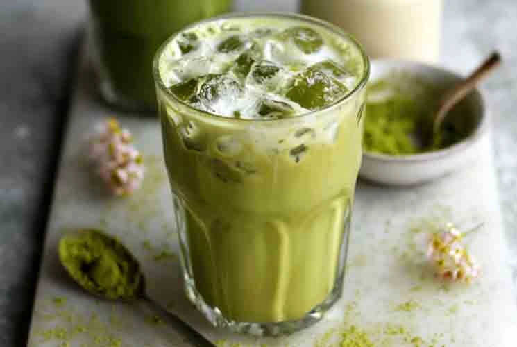 Glass of iced matcha latte made with almond milk and matcha powder, served on a white board with matcha spoon and spring flowers.