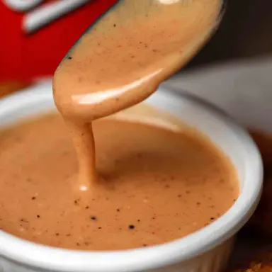 Close-up of a spoonful of creamy, pepper-speckled Raising Cane’s Sauce dripping into a white ramekin, served with crispy chicken tenders.