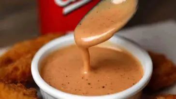 A spoon drizzling creamy Raising Cane’s Sauce into a white ramekin, surrounded by golden chicken tenders, with a red Cane’s cup in the background.