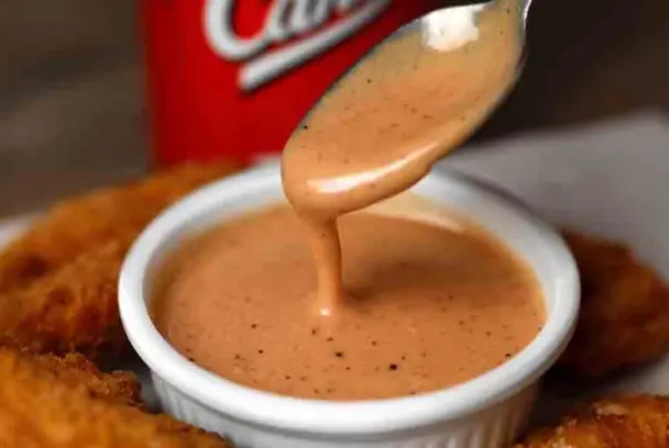 A spoon drizzling creamy Raising Cane’s Sauce into a white ramekin, surrounded by golden chicken tenders, with a red Cane’s cup in the background.