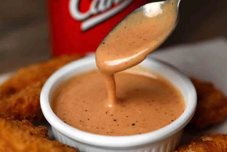 A spoon drizzling creamy Raising Cane’s Sauce into a white ramekin, surrounded by golden chicken tenders, with a red Cane’s cup in the background.