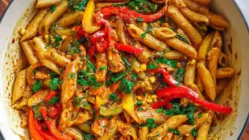 Close-up of Rasta Pasta in a white pot, featuring penne pasta mixed with sliced red, yellow, and green bell peppers, seasoned jerk chicken, and fresh herbs in a creamy sauce.