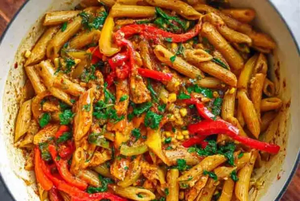 Close-up of Rasta Pasta in a white pot, featuring penne pasta mixed with sliced red, yellow, and green bell peppers, seasoned jerk chicken, and fresh herbs in a creamy sauce.
