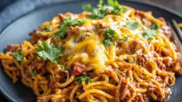 Plate of slow cooker spaghetti casserole topped with melted cheese, ground beef, and fresh parsley, served on a dark plate with a fork on the side.