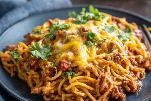 Plate of slow cooker spaghetti casserole topped with melted cheese, ground beef, and fresh parsley, served on a dark plate with a fork on the side.