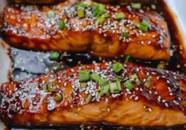 Baked teriyaki salmon fillets glazed with a rich sweet-savory sauce, topped with sesame seeds and chopped green onions, served in a baking dish.